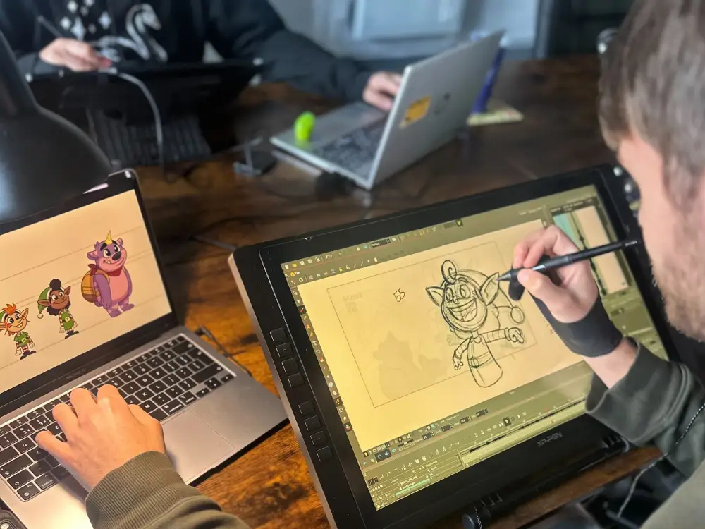 A behind the scenes photo of Spirit Bridge Studio animator, Nathan J. Hill, hand drawing Tappy the elf for the "Smile for Us" music animation on a large graphics tablet. A laptop beside him shows the coloured character art.