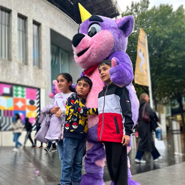 A heartwarming photo of the Bucky the Unicorn Bear mascot giving a big, friendly hug to a group of smiling children at the Bullring shopping centre in Birmingham. The scene is full of joy and community spirit.