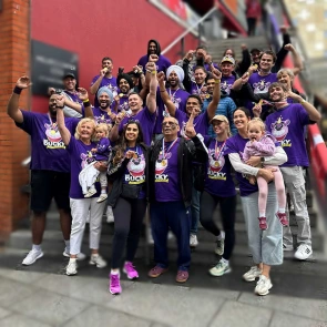 A joyous group photo of 'Team Bucky' celebrating their fundraising success at the Dragon Boat Race. The team members, all wearing matching purple Bucky t-shirts and gold medals, are cheering with their arms in the air on an outdoor staircase.