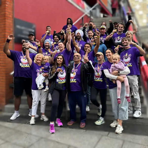 A joyous group photo of 'Team Bucky' celebrating their fundraising success at the Dragon Boat Race. The team members, all wearing matching purple Bucky t-shirts and gold medals, are cheering with their arms in the air on an outdoor staircase.