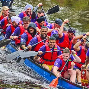 An action photo of 'Team Bucky' (Spirit Bridge Studio) paddling intensely in their blue dragon boat during their third race. Team members, wearing purple Bucky t-shirts and red life vests, are focused and working together, splashing water as they race.
