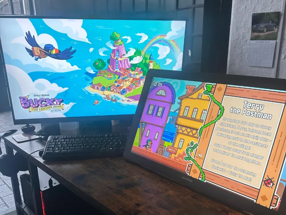 A behind the scenes photo at Spirit Bridge Studio, showing the 'Terry the Postbird' pixel game description on a drawing tablet, with Sunnyside Island concept art on the monitor.